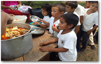 FMI rollover image of young Honduran boy receiving a hot school lunch then second image is a group of Honduran school boys sitting at a plastic table eating a hot school lunch.